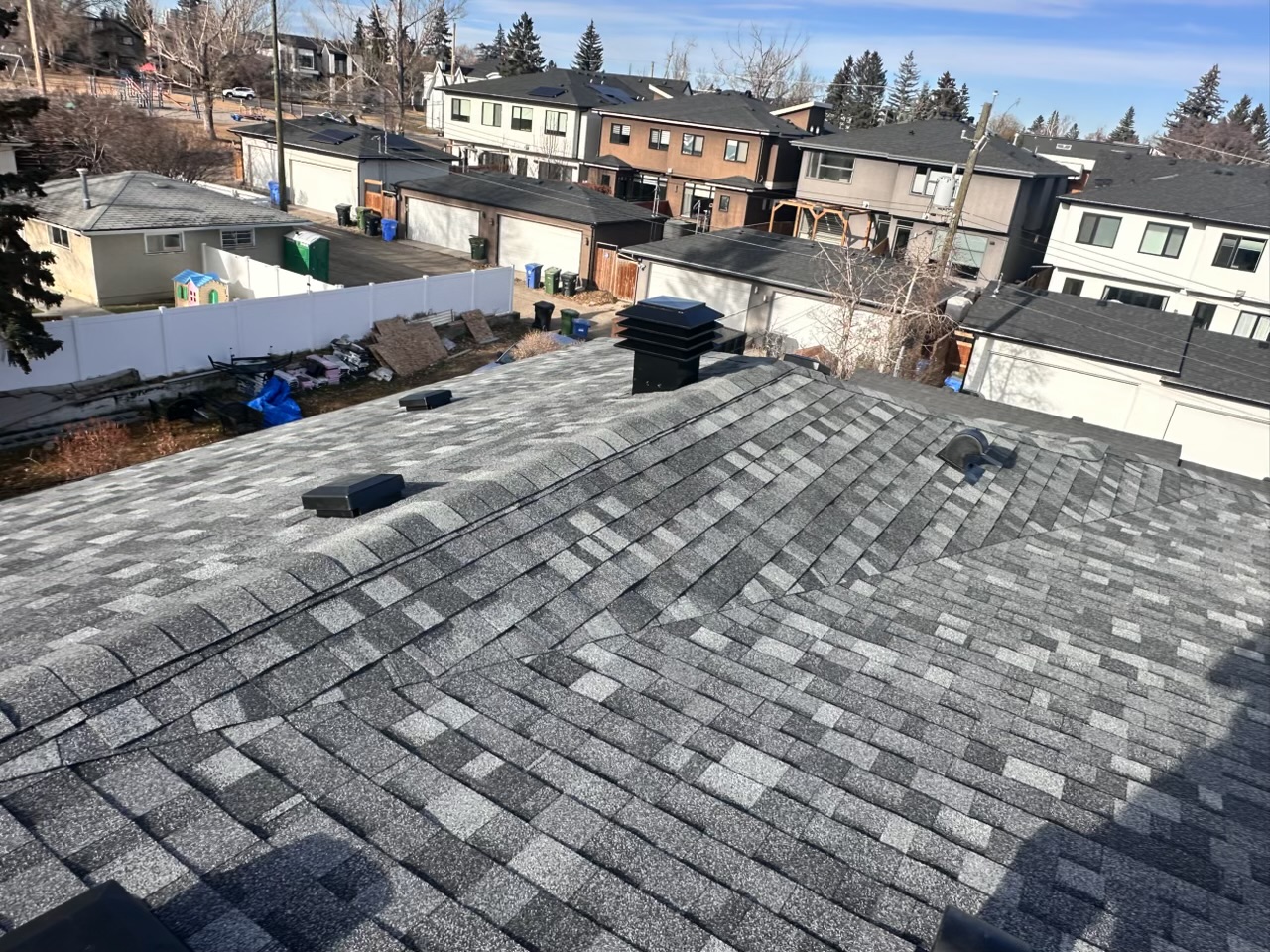 Grey shingle roof with ridge vents and stack flashing, seen from the peak toward the back lane
