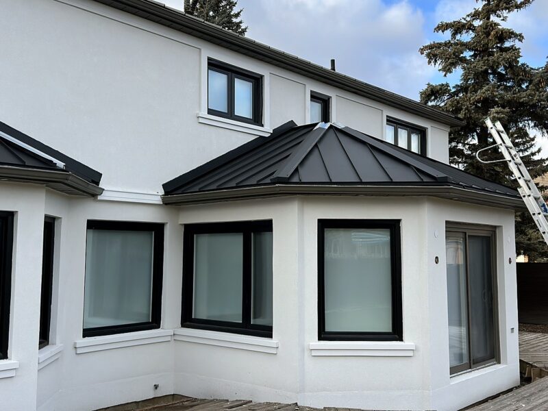 Two-storey stucco house with black standing seam metal roof accents over the bay windows, winter