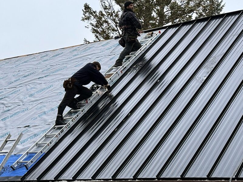 Roofing crew installing black standing seam metal panels over underlayment in winter